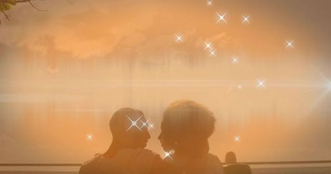 Couple watching sunset on waterfront bench leaning together in golden silhouette