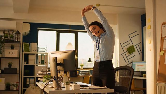 Office worker stretching at desk wearing headset and id badge in modern bright workspace