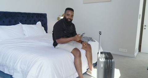 Man Relaxing on Hotel Bed with Smartphone and Luggage