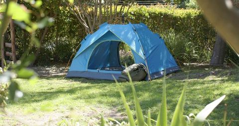 Blue camping tent sitting on sunny backyard lawn with duffel bag and leafy shade near trees