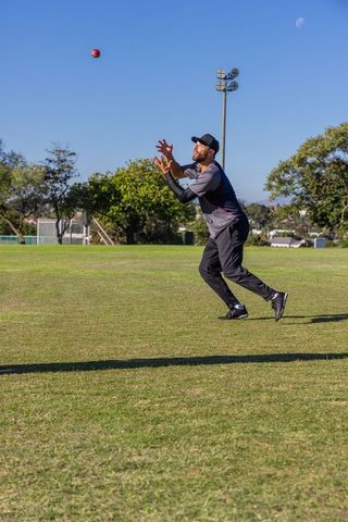 Athlete running and reaching to catch red cricket ball on grassy field under blue sky