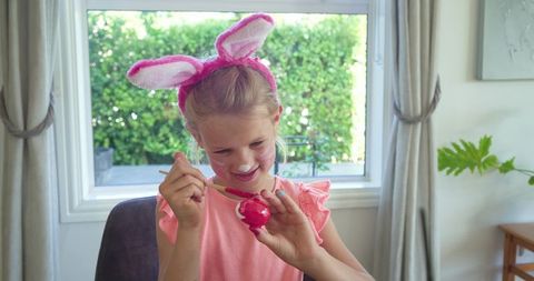 Young girl painting bright pink easter egg while wearing bunny ear headband in sunlit room