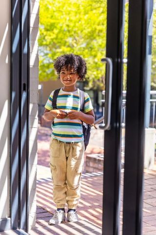 African american child standing in sunlit school doorway carrying backpack and smiling