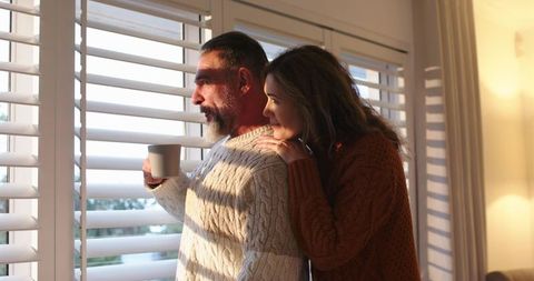 Middle-aged couple enjoying cozy morning by window with plantation shutters and warm sweaters