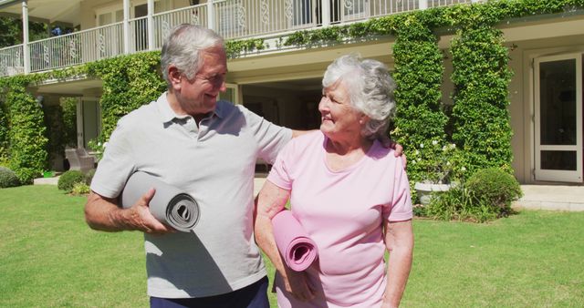 Happy caucasian senior couple exercising embracing walking carrying yoga mats in garden in the sun. at home in isolation during quarantine lockdown.