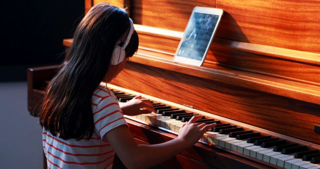 Young girl practicing piano using headphones and a digital tablet for guidance. Perfect for depicting online music education, child-focused learning techniques, and the integration of technology in musical practice. Ideal for blogs, educational websites, and advertisements related to e-learning platforms for children.