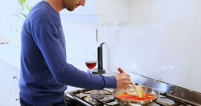 Man preparing pasta sauce on stove in bright, modern kitchen. Ideal for domestic life themes, cooking tutorials, home culinary skills, kitchen appliance promotions, food blog visuals.