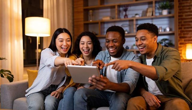 Group of friends laughing and pointing at tablet while relaxing on cozy sofa