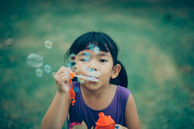 Young girl with black hair, standing on grassy area, enjoying blowing soap bubbles. Ideal for using in themes related to childhood, outdoor play, summer activities, joy, and carefree moments. Use in educational materials, advertisements for children's products, or blog posts about parenting and children's activities.
