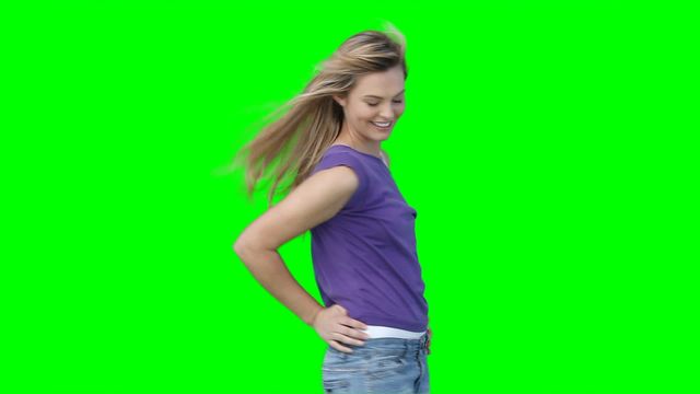 Young woman standing with a hand on her hip, smiling while wind blows her hair. Dressed in casual clothes including a purple top and jeans. Suitable for use in advertising, fashion shoots, or background replacement projects.