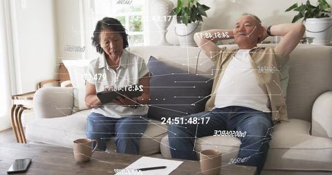 Chinese couple relaxing on sofa using tablet with hud overlays in cozy living room
