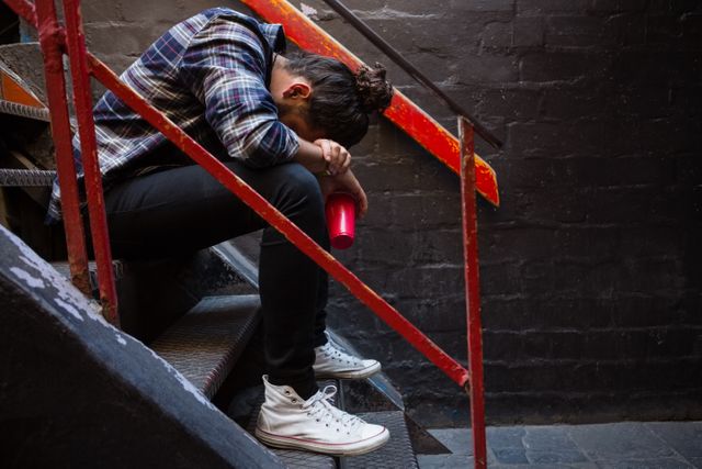 Man sitting on staircase in bar, appearing drunk and distressed. Useful for illustrating themes of alcohol abuse, mental health struggles, loneliness, and urban nightlife. Can be used in articles, blogs, and campaigns related to addiction, depression, and social issues.
