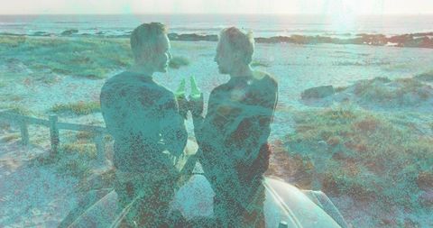 Two men clinking bottles while sitting on car roof overlooking sunset coastline