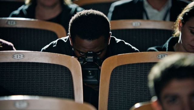 Cameraman peering through viewfinder in dim auditorium capturing live event