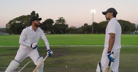 Two cricketers practicing batting on worn club pitch at dusk under floodlights