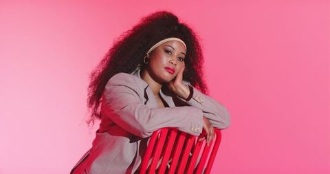 Mid 30s african american woman leaning on red chair wearing blazer and headband on pink background