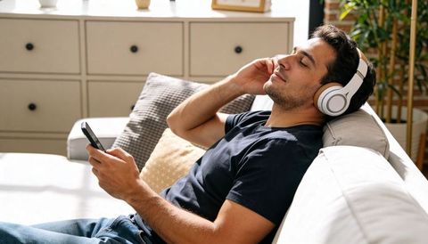 Man relaxing on couch listening to headphones and scrolling smartphone in bright living room