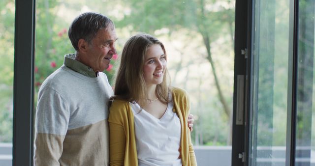 Smiling senior caucasian father and teenage daughter embracing and looking out of window. family spending time at home.