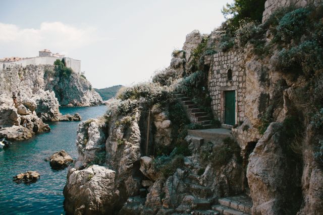 Stone structure nestled on a rocky Mediterranean coast with steps leading to a small door. Turquoise water and scenic cliffs enhance the beauty. Ideal for illustrating travel destinations, architectural design, serene retreats, and landscapes.