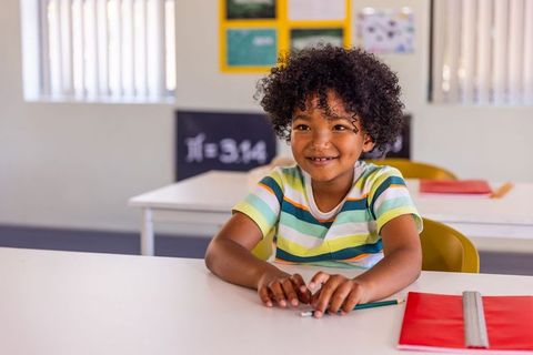 Smiling schoolboy sitting at classroom desk holding pencil and red notebook