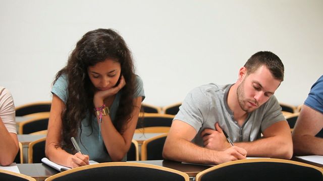 Students sitting at desks, focused on taking notes during a classroom lecture. Ideal for content related to education, learning environments, student life, academic tutorials, and university promotional materials.