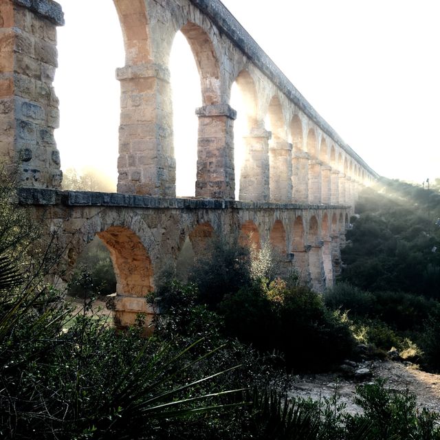 This ancient stone aqueduct with sunlight streaming through the arches highlights its detailed stonework and architectural significance. Ideal for use in travel guides, history textbooks, architectural studies, and promotional materials for historical landmarks.