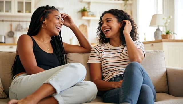 Two friends laughing on couch in bright open kitchen, cozy casual home conversation