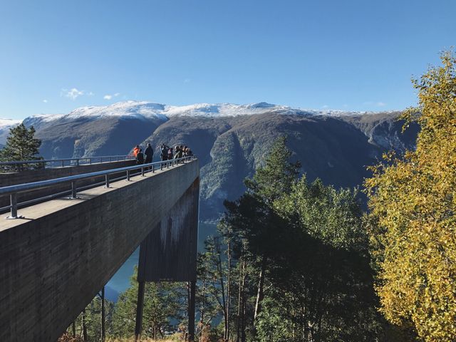 A group of tourists stand on an architectural overlook bridge against a backdrop of mountains with a blue sky overhead. This is a suitable image for travel brochures, outdoor adventure websites, or promotional content for scenic locations. The vibrant natural setting conveys the beauty of rural and mountain travel experiences.