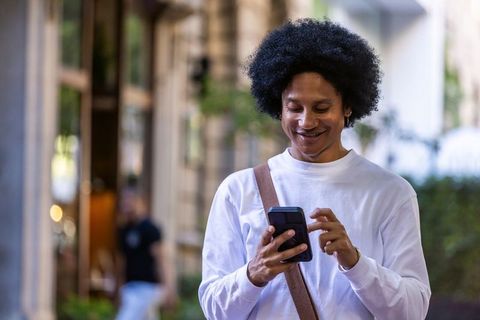 African american commuter smiling using smartphone on city sidewalk with crossbody bag
