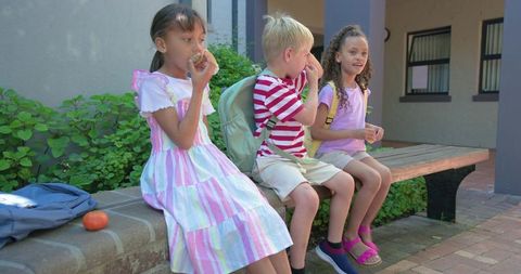 Diverse schoolchildren sitting on courtyard bench eating snacks with backpacks