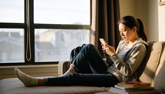 Sunlit student lounging on sofa tapping smartphone in cozy urban apartment, relaxed study