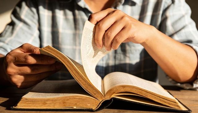 Man turning pages of vintage book in sunlit reading nook, hands and spine detail