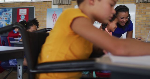 Biracial schoolboy in wheelchair, sitting in classroom making notes, teacher in background. children in primary school.