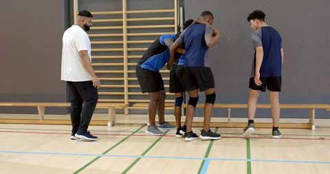 Diverse male team supporting injured teammate during indoor training, coach observing