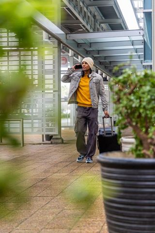 Urban traveler walking with rolling suitcase talking on smartphone under glass canopy