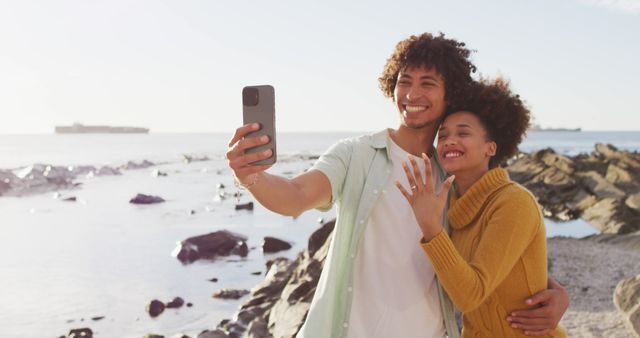 African american couple taking a selfie and showing their ring standing on the rocks near the sea. love and relationship concept