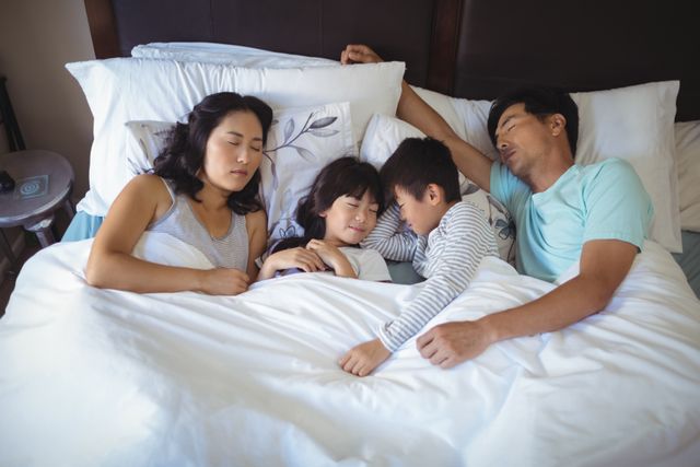 Asian family of four peacefully sleeping together in bed, showcasing bonding and togetherness. Ideal for use in parenting articles, family health and wellness blogs, home comfort advertisements, or family-oriented product promotions.