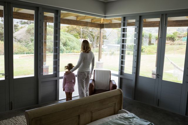 Mother and daughter standing together looking out the window, creating a warm and intimate family moment. Ideal for use in parenting blogs, family lifestyle articles, home decor inspiration, and advertisements promoting family-oriented products.