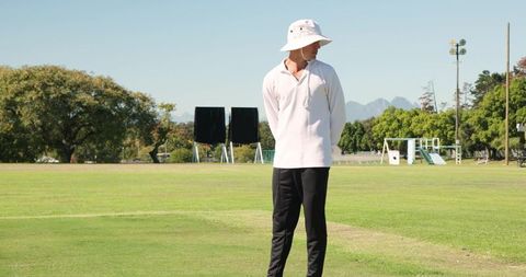 Match official standing on green sports field wearing bucket hat and relaxed white shirt