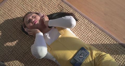 Mid adult asian woman lying on sunlit rug listening with headphones in yellow overalls