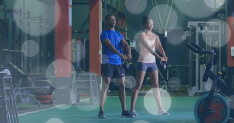 Dual trainer kettlebell workout on gym turf with spin bike and weight racks, partners