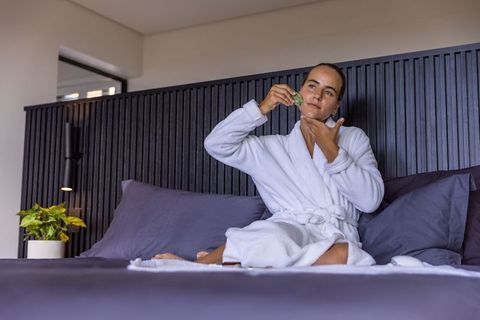 Woman using jade facial roller while relaxing in white bathrobe on bed