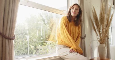 Mid adult woman sitting on windowsill wearing mustard sweater in sunlit living room