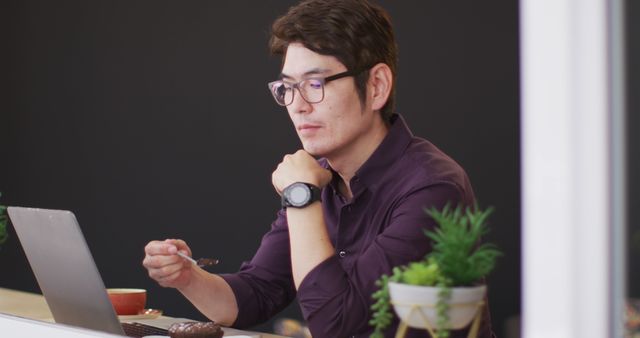 Adult man wearing glasses and casual clothing, working on a laptop at home, while holding a spoon and having a snack. Great for themes involving remote work, modern lifestyle, technology, concentration, and home office setups. Suitable for use in blogs, online articles, work-from-home guides, and lifestyle magazines.