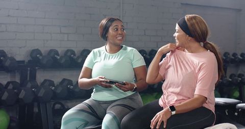 Diverse women chatting on gym bench with tablet and dumbbells, fitness friendship