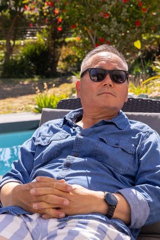 Mature asian man relaxing poolside on lounge chair wearing denim shirt and sunglasses