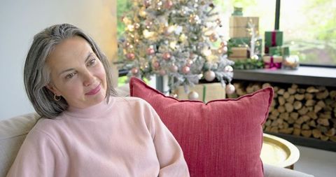 Mature woman relaxing on sofa with red pillow near frosted Christmas tree and gifts