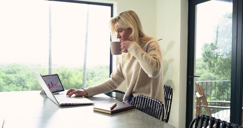 Woman sipping coffee while checking laptop charts in remote work at sunlit kitchen counter
