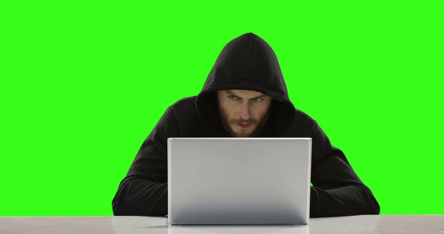 A Caucasian man in a hooded sweatshirt is intently using a laptop against a green screen background, with copy space. His focused demeanor and attire suggest he could be portraying a hacker or a tech enthusiast in a simulated environment.