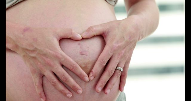 A close-up of a pregnant woman holding her belly with hands forming a heart shape. There is a wedding ring on her finger, signifying a family bond. This image is ideal for use in content related to pregnancy, motherhood, bonding, prenatal care, and family planning. It can be used on websites, blogs, social media, and advertisements focusing on women's health and maternity care.